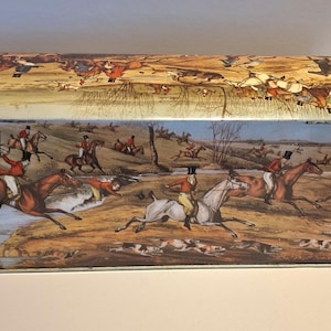 Puede incluir: Lata metálica rectangular con una ilustración de escena de caza. La tapa y los lados presentan jinetes a caballo, perros y un paisaje con árboles y un río. La paleta de colores incluye marrones, rojos, azules y verdes.
