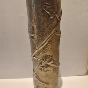 May include: A tall, cylindrical brass vase with a textured, hammered surface. The exterior features a raised, embossed design of winding vines and leaves that wrap around the body. The top rim has a distinct, wavy, scalloped edge finish.