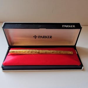 Pode incluir: Uma caneta-tinteiro Parker dourada em uma caixa aberta preta e branca com interior de veludo vermelho. A caneta tem um design texturizado e é exibida em uma almofada de veludo vermelho. O logotipo da Parker está impresso na caixa.