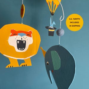 May include: A hanging mobile featuring a yellow lion with an open mouth, a blue elephant with a party hat, a yellow hot air balloon with orange stars, and a blue rabbit emerging from a black top hat, all suspended from a white metal frame.