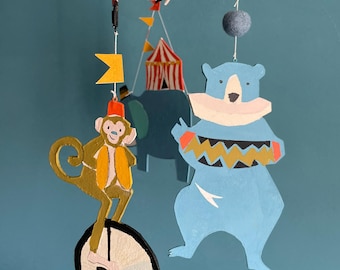 May include: A whimsical circus-themed mobile featuring a monkey on a unicycle, a blue bear, and a circus tent. The mobile is suspended by strings and includes colorful flags and a rabbit. The figures are painted in bright colors against a teal background.