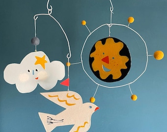 May include: A whimsical baby mobile featuring a white cloud with a smiling face and star, a white bird with yellow accents, and a sun with a smiling face. The mobile is suspended by a white wire frame with yellow and gray accents.