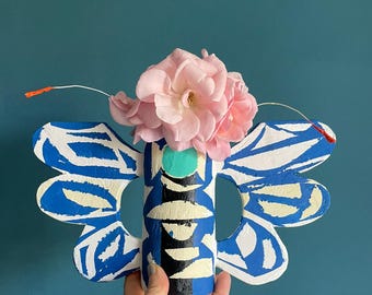 May include: A whimsical, hand-painted butterfly-shaped vase in blue, white, and black, holding pink roses. The vase has a cylindrical body and stylized wings. The design features geometric patterns and a turquoise accent.