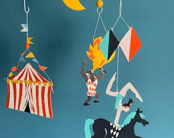 May include: A whimsical mobile featuring circus-themed cutouts. Includes a yellow crescent moon, a circus tent with red and white stripes, and performers. The performers include a fire-eater, a woman doing a handstand on a horse, and geometric shapes.
