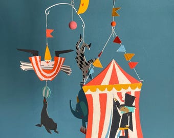 May include: A whimsical mobile featuring circus-themed cutouts. The mobile includes a yellow crescent moon, a clown, a seal balancing a ball, a zebra, an elephant, and a circus tent with a performer. The colors are red, orange, blue, and black.