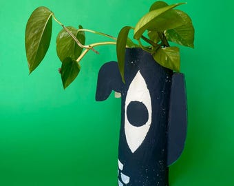 May include: A handmade, black and white painted vase with a whimsical face design, holding green leafy plants. The vase has a cylindrical shape with a large eye and ear details. The background is a solid green.