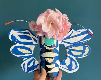 May include: A butterfly-shaped decorative object featuring a central cylindrical body painted with blue, black, white, and teal patterns. Large blue and white wings extend from the sides. A cluster of light pink flowers sits atop the center.