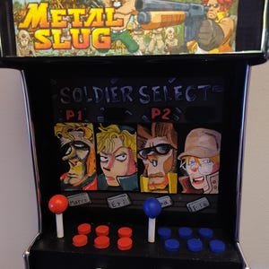 Peut inclure: Miniature borne d'arcade avec le jeu "Metal Slug". Le meuble noir présente un auvent coloré et un écran "Soldier Select". Il comprend des joysticks et des boutons rouges et bleus, avec des portraits de personnages en dessous.