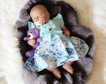 May include: A sleeping baby doll wearing a blue and white floral dress with a light blue cardigan and a pacifier. The doll is holding a purple teddy bear.