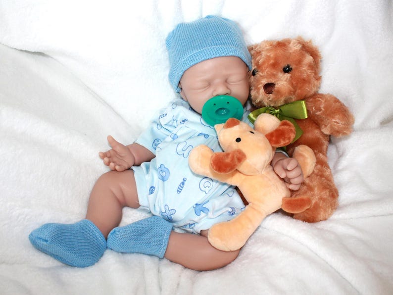 A baby doll with a pacifier, accompanied by a soft stuffed animal, resting on a light-colored blanket.