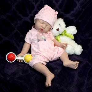 A baby doll holding a pacifier, next to a plush stuffed animal, set against a pastel background.
