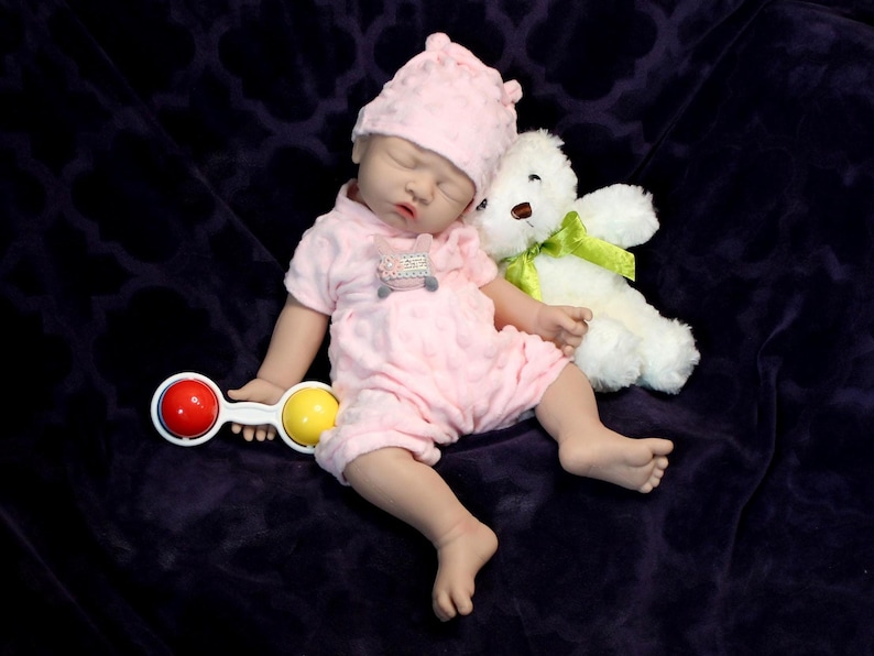 A baby doll holding a pacifier, next to a plush stuffed animal, set against a pastel background.