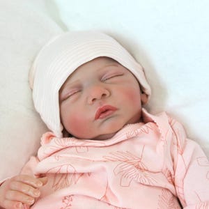 May include: A sleeping baby doll wearing a pink and white outfit with a white knit hat. The doll has closed eyes and rosy cheeks.