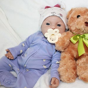 May include: A baby doll wearing a light blue onesie and a white hat with a red bow. The doll has a pacifier and is next to a brown teddy bear with a green ribbon.