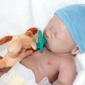 A baby doll with a pacifier, accompanied by a soft stuffed animal, resting on a light-colored blanket.