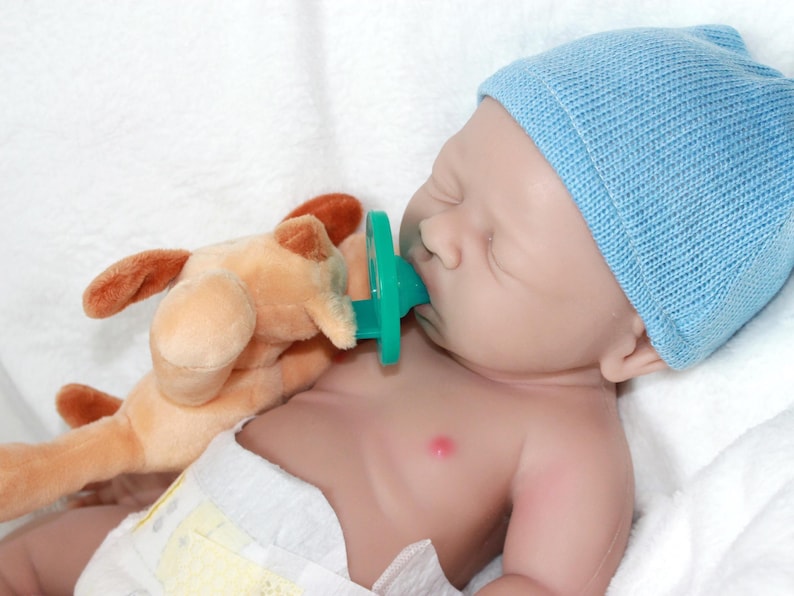 A baby doll with a pacifier, accompanied by a soft stuffed animal, resting on a light-colored blanket.