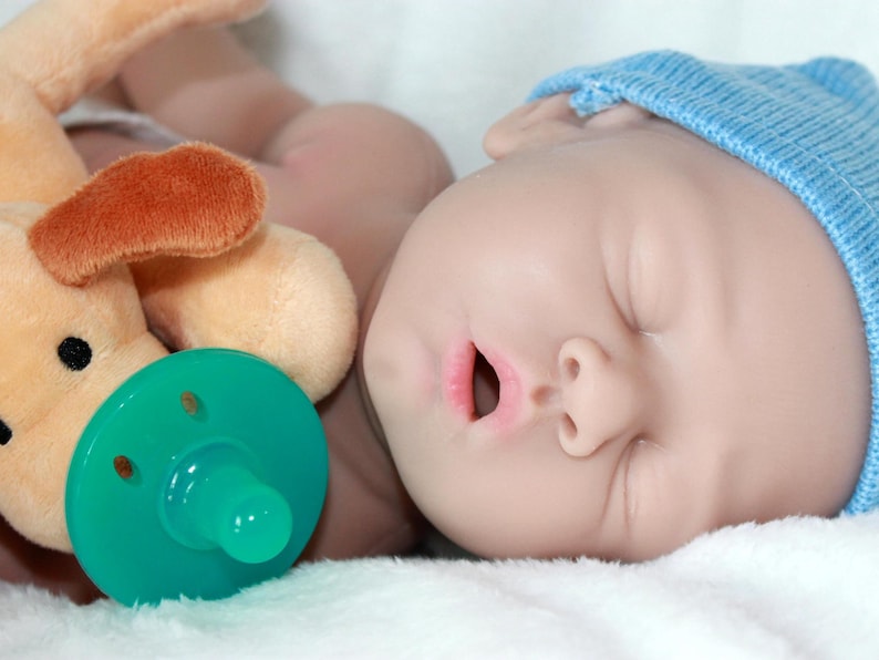 A baby doll with a pacifier, accompanied by a soft stuffed animal, resting on a light-colored blanket.