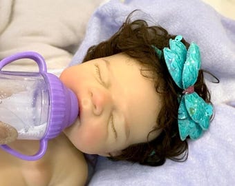 May include: A lifelike baby doll with closed eyes, being fed from a purple bottle. The doll has dark brown hair and a teal bow. The doll is lying on a light purple blanket.