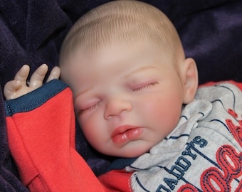 May include: A realistic baby doll with closed eyes, rosy cheeks, and a slightly open mouth. The doll wears a red and gray baseball-themed outfit with the text "Daddy's". The doll is resting on a dark blue blanket.