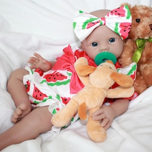 Op de afbeelding: Een babypop die een watermeloenprintjurk en een hoofdband met een strik draagt. De pop houdt een pluche hond en een teddybeer vast. De pop heeft blauwe ogen en draagt een speen.