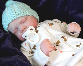 Puede incluir: Una muñeca bebé realista con un gorro de punto azul claro con un pompón y un chupete blanco. El bebé viste un body blanco con un estampado repetido de perros marrones. La muñeca está acostada con los ojos cerrados.
