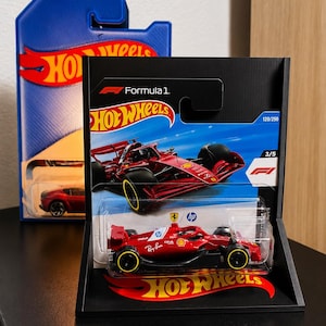 Könnte beinhalten: Ein rotes Hot Wheels Formel-1-Modellauto mit schwarzen Akzenten und Logos von HP, Ray-Ban und Shell. Das Auto befindet sich in der Originalverpackung, die eine blau-weiße Karte mit einer Abbildung des Wagens und dem Schriftzug Formula 1 enthält.