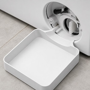 Può includere: Una vaschetta di scarico in plastica bianca fissata sul pannello frontale inferiore di una lavatrice. La vaschetta è aperta e rivela un foro di accesso circolare con una guarnizione in gomma nera e un filtro interno bianco visibile.