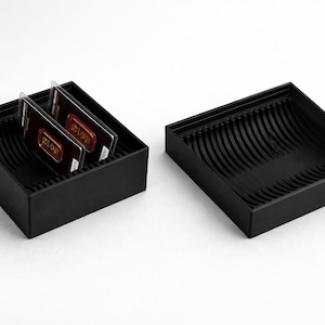 Pode incluir: Duas bandejas retangulares pretas com interior canelado. Uma bandeja contém dois cartões retangulares. As bandejas estão sobre uma superfície branca. Os cartões têm um design dourado e vermelho.