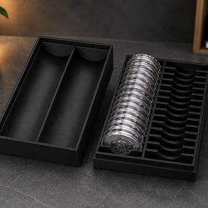 Könnte beinhalten: Eine schwarze, rechteckige Aufbewahrungsbox mit zwei offenen Fächern und einem separaten schwarzen Einsatz mit einer Reihe kreisförmiger Aussparungen. Der Einsatz enthält einen Stapel runder, transparenter Münzkapseln.