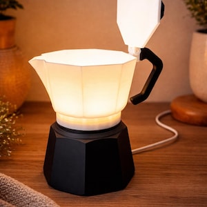 Può includere: Lampada a forma di caffettiera bianca e nera con interno luminoso. La lampada ha una base nera, una parte superiore bianca e un manico nero. La lampada è collegata a una presa a muro con un cavo bianco. La lampada è su una superficie di legno.