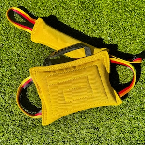 May include: A yellow rectangular fabric dog toy featuring a central stitched panel and a black plastic handle. The toy includes integrated straps with red, yellow, and black stripes, designed with a durable textured surface for interactive play.