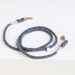 May include: A grey dog lead made from thick, twisted rope. It features gold-coloured metal clasps and a small, round charm. The ends of the lead are accented with a lighter grey colour.