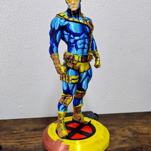 May include: A standing figurine of a comic book character with brown hair, wearing a blue suit with yellow accents, a yellow utility belt, and brown boots. The character has a yellow visor over the eyes and stands on a circular yellow and red base.