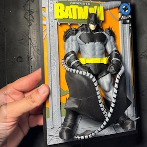 Könnte beinhalten: Ein 3D-Wandkunstwerk mit einer skulptierten Batman-Figur in einem grau-schwarzen Anzug mit gelbem Gürtel. Die Figur ist vor einem orangefarbenen Hintergrund mit grauem Rahmen platziert. Der Text ABSOLUTE BATMAN und ALL IN ist zu sehen.