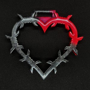 May include: A heart-shaped pendant with a barbed wire design. The top portion is red and the bottom is silver. The pendant has a small rectangular loop at the top for hanging. The background is black.