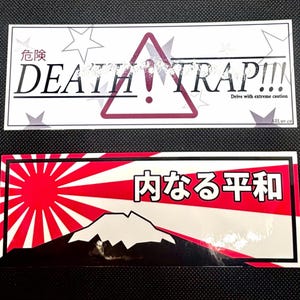 Puede incluir: Dos pegatinas rectangulares. La superior muestra "DEATH TRAP!!!" con un símbolo de advertencia y texto japonés. La inferior presenta un diseño de sol rojo con una silueta de montaña y texto japonés.