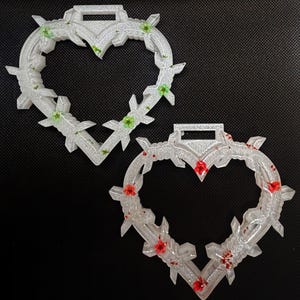 May include: Two heart-shaped pendants with a barbed wire design. One pendant features green flowers, the other red. Both are clear with glitter and a rectangular loop at the top for hanging. The pendants are on a dark background.