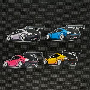 May include: Four colorful car stickers. Each sticker features a stylized sports car with a large rear wing. The cars are in shades of gray, blue, pink, and yellow, with black and white accents. The stickers are on a black background.