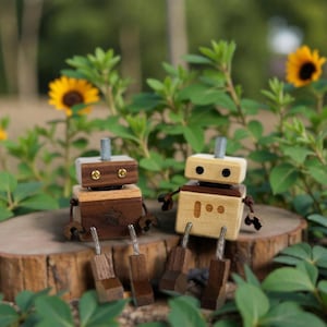 May include: Two wooden robot figurines with square heads, bodies, and legs, sitting on a tree stump. One robot is light-colored wood, the other is dark. Both have metal antennae and small arms. The background includes greenery and sunflowers.