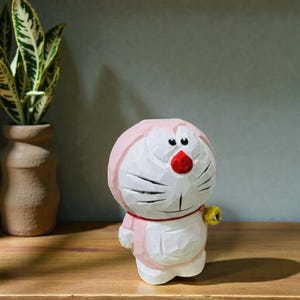 May include: A carved wooden figurine of a cartoon character with a white body, pink accents, and a red nose. The figure has black eyes and a yellow bell on a red collar. The figurine is on a wooden surface.