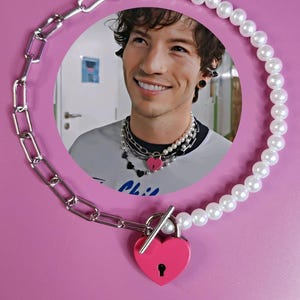 Puede incluir: Un collar con un candado rosa en forma de corazón, una cadena plateada y un collar de perlas. La imagen también muestra a una persona que lleva el collar. El fondo es de color rosa liso.
