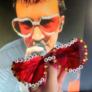 Peut inclure: Lunettes de soleil rouges avec les mots "TWENTY ONE PILOTS TOUR CLANCY" épelés en perles blanches. Les verres sont d'un rouge vif et les montures sont décorées de strass jaunes et rouges. En arrière-plan, une personne.