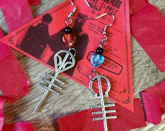 May include: Silver-toned earrings featuring a stylized symbol with a cross-like design. Each earring has a heart-shaped bead in either red or blue, and a black bead. The earrings are displayed on a red background with text.