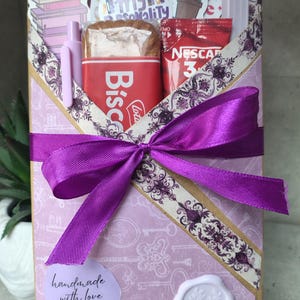 Op de afbeelding: Een cadeauverpakking in lavendelkleurig papier met een patroon, vastgebonden met een paars satijnen lint. Het bevat een pen, snacks en koffie. De doos is versierd met stickers en een label met "handmade with love".