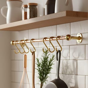 May include: A kitchen scene with a copper-colored hanging rod and gold-colored hooks. Various kitchen utensils hang from the hooks, including a whisk, a wooden spoon, and a black pan. A sprig of rosemary and a mug are also visible.