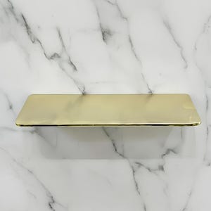 Może przedstawiać: Prostokątna, złota półka. Półka wykonana jest z błyszczącego, metalicznego materiału i zamontowana na białej marmurowej ścianie z szarymi żyłkami. Półka ma prosty, nowoczesny design.