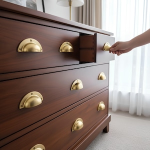 Puede incluir: Una cómoda de madera marrón oscuro con tiradores de cajón en forma de media luna de color latón. Un cajón está parcialmente abierto, revelando el interior. La cómoda tiene un diseño clásico y está en una habitación bien iluminada.