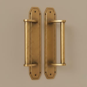 May include: Two antique-style door pulls with a gold finish. Each pull has a rectangular backplate with rounded edges and a cylindrical handle. The hardware is mounted with small screws, and the overall design is vintage.