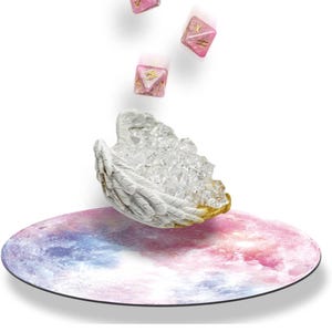 May include: A fantasy-themed still life featuring three pink and gold dice with symbols, a white angel wing-shaped crystal holder with gold accents, and a round mat with a colorful galaxy design.