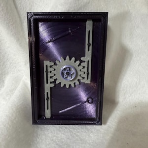 May include: A rectangular, 3D-printed puzzle with a dark purple interior and a black frame. The puzzle features a central gear with a star emblem, flanked by two gray, linear components. The design suggests a mechanical or industrial theme.